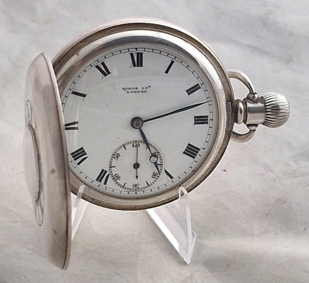 A polished silver half hunter pocket watch displaying an engraved outer ring with black Roman numerals surrounding a central circular glass window. View II