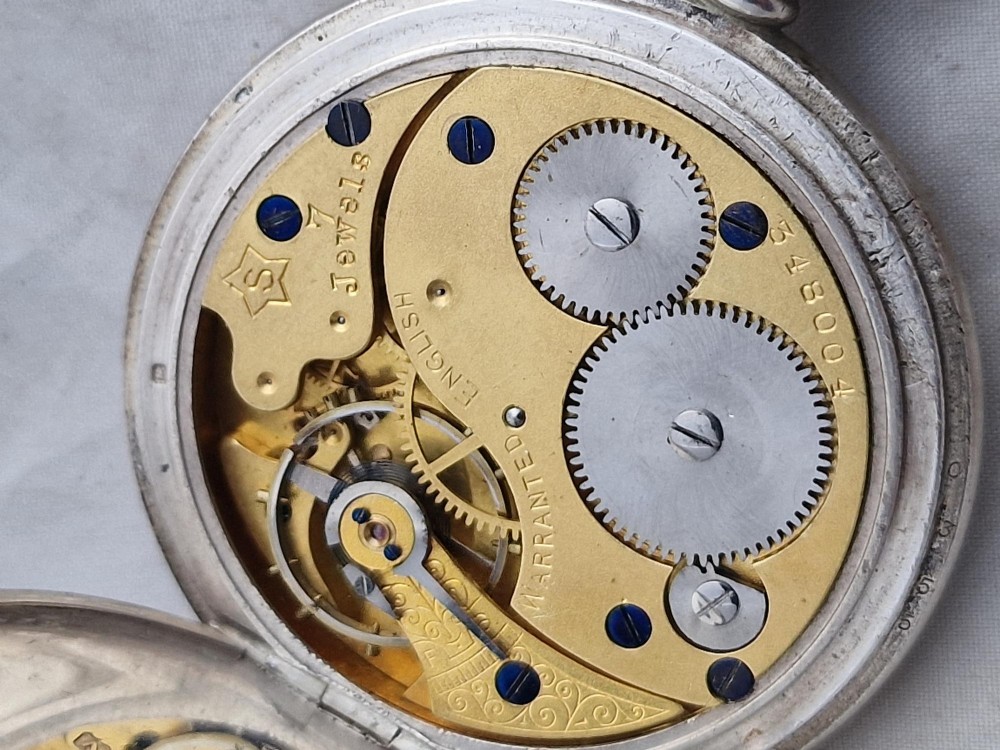 A polished silver half hunter pocket watch displaying an engraved outer ring with black Roman numerals surrounding a central circular glass window. View IV