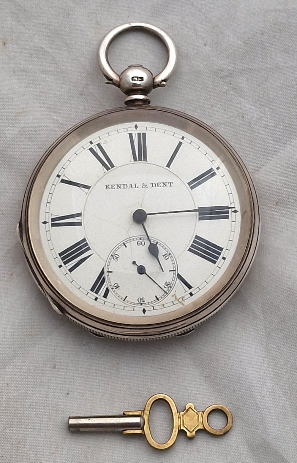 An open-face silver pocket watch displaying a white enamel dial with black Roman numerals, a subsidiary seconds dial, and an accompanying brass winding key.