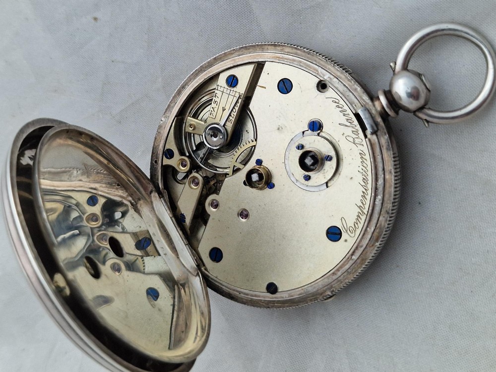An open-face silver pocket watch displaying a white enamel dial with black Roman numerals, a subsidiary seconds dial, and an accompanying brass winding key. View III