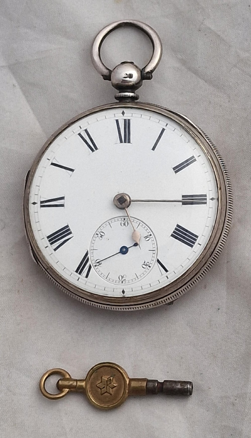 An antique open-face silver pocket watch featuring a white enamel dial with Roman numerals, a subsidiary seconds dial, and an accompanying brass winding key.