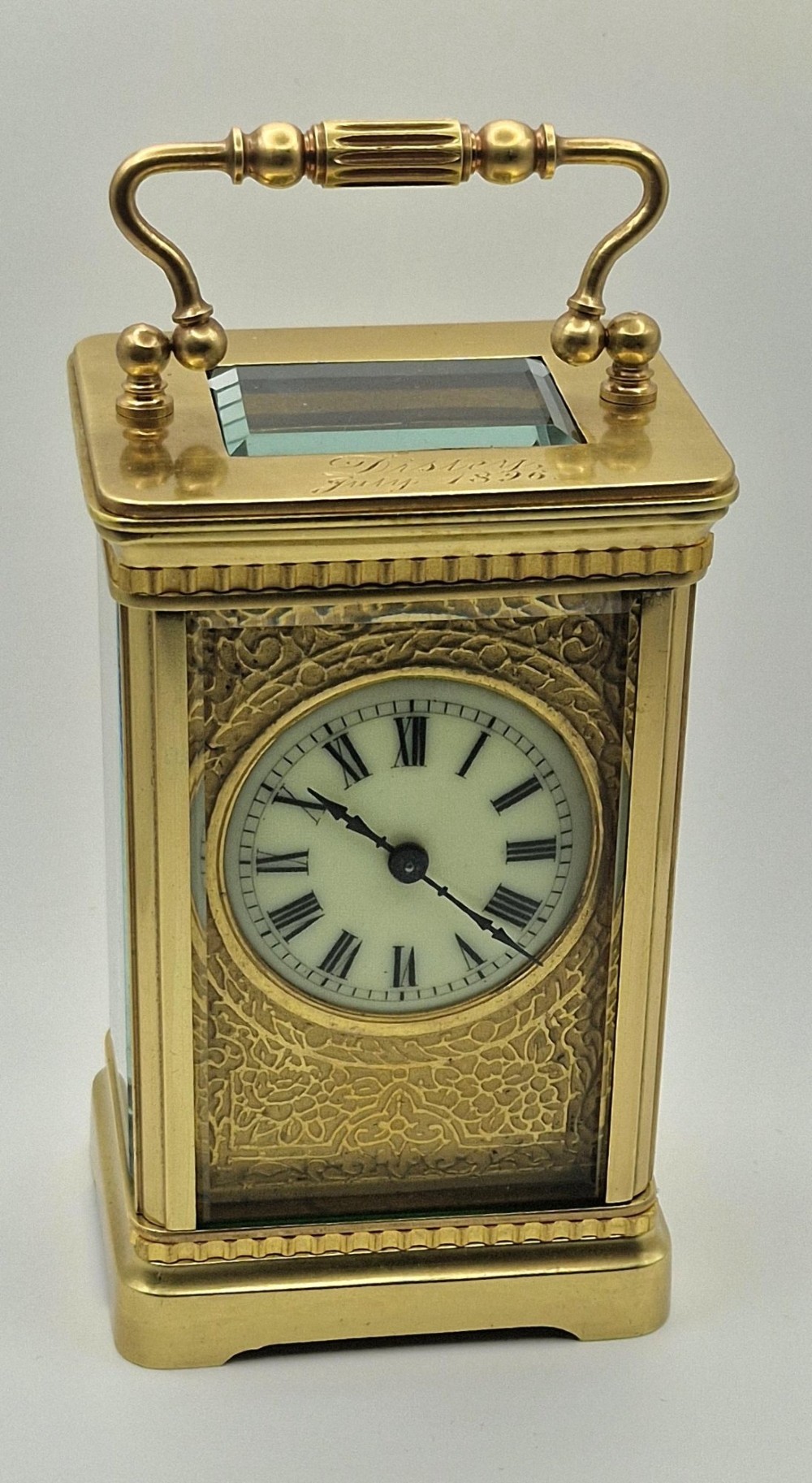 An antique brass carriage clock with an engraved gilded dial plate, white enamel face, and carrying handle, set next to its fitted brown leather transport case. View II