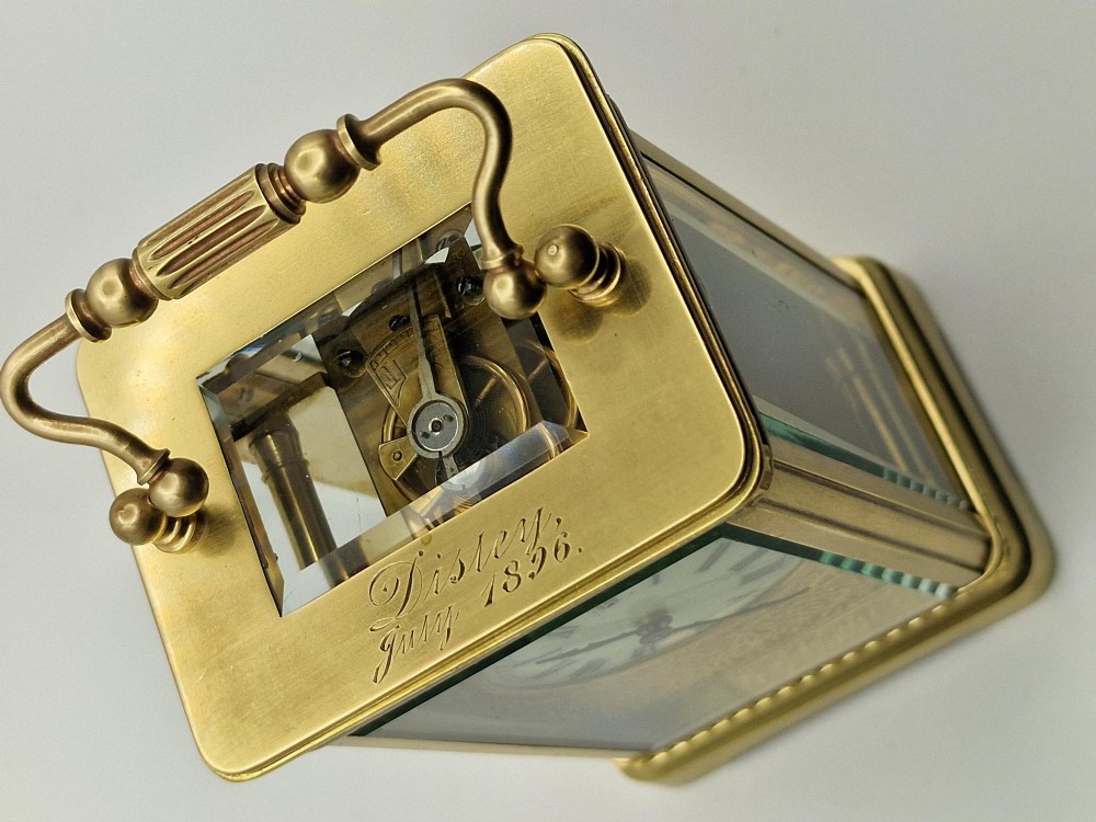 An antique brass carriage clock with an engraved gilded dial plate, white enamel face, and carrying handle, set next to its fitted brown leather transport case. View V