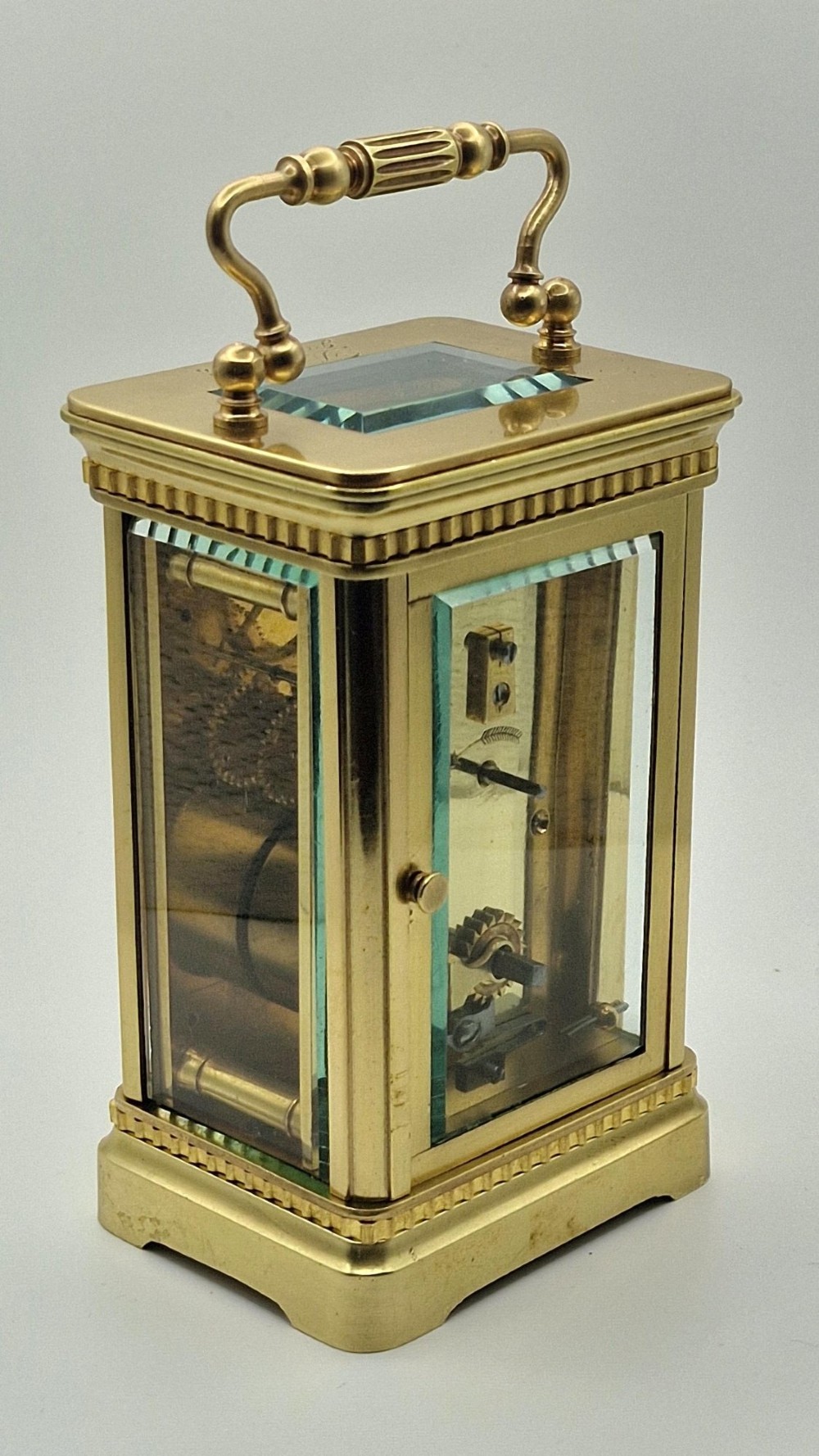An antique brass carriage clock with an engraved gilded dial plate, white enamel face, and carrying handle, set next to its fitted brown leather transport case. View VI