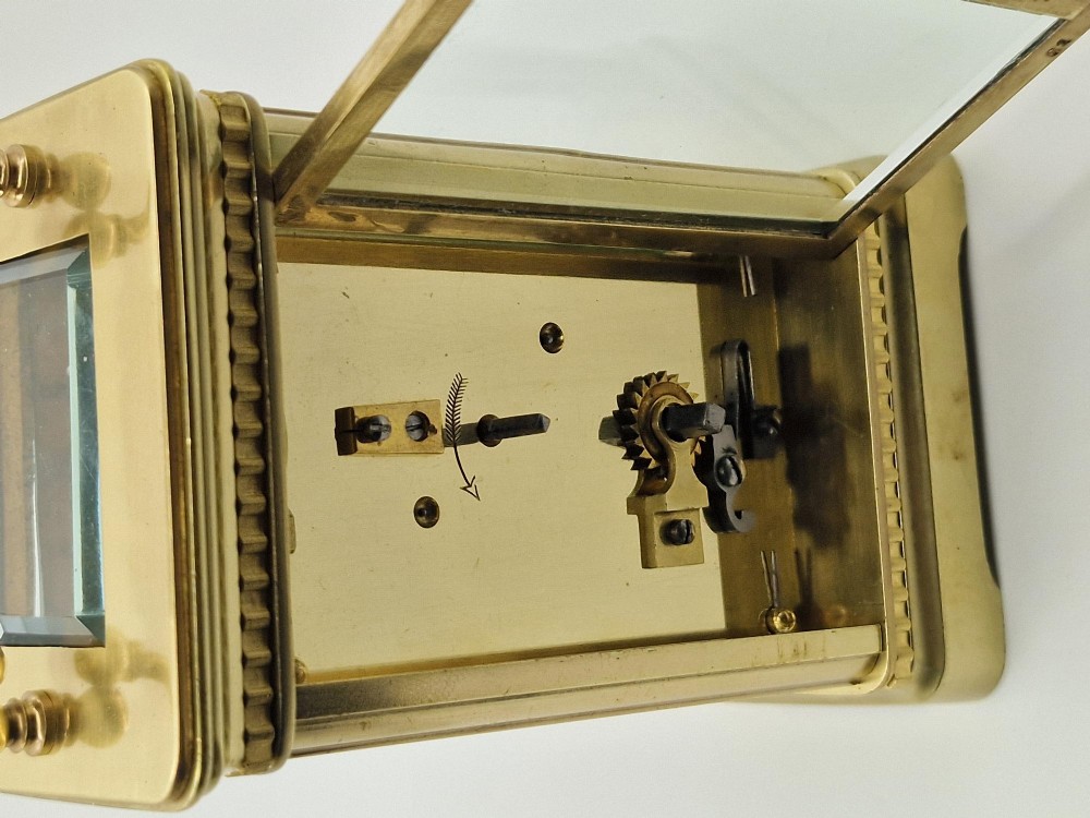 An antique brass carriage clock with an engraved gilded dial plate, white enamel face, and carrying handle, set next to its fitted brown leather transport case. View VII