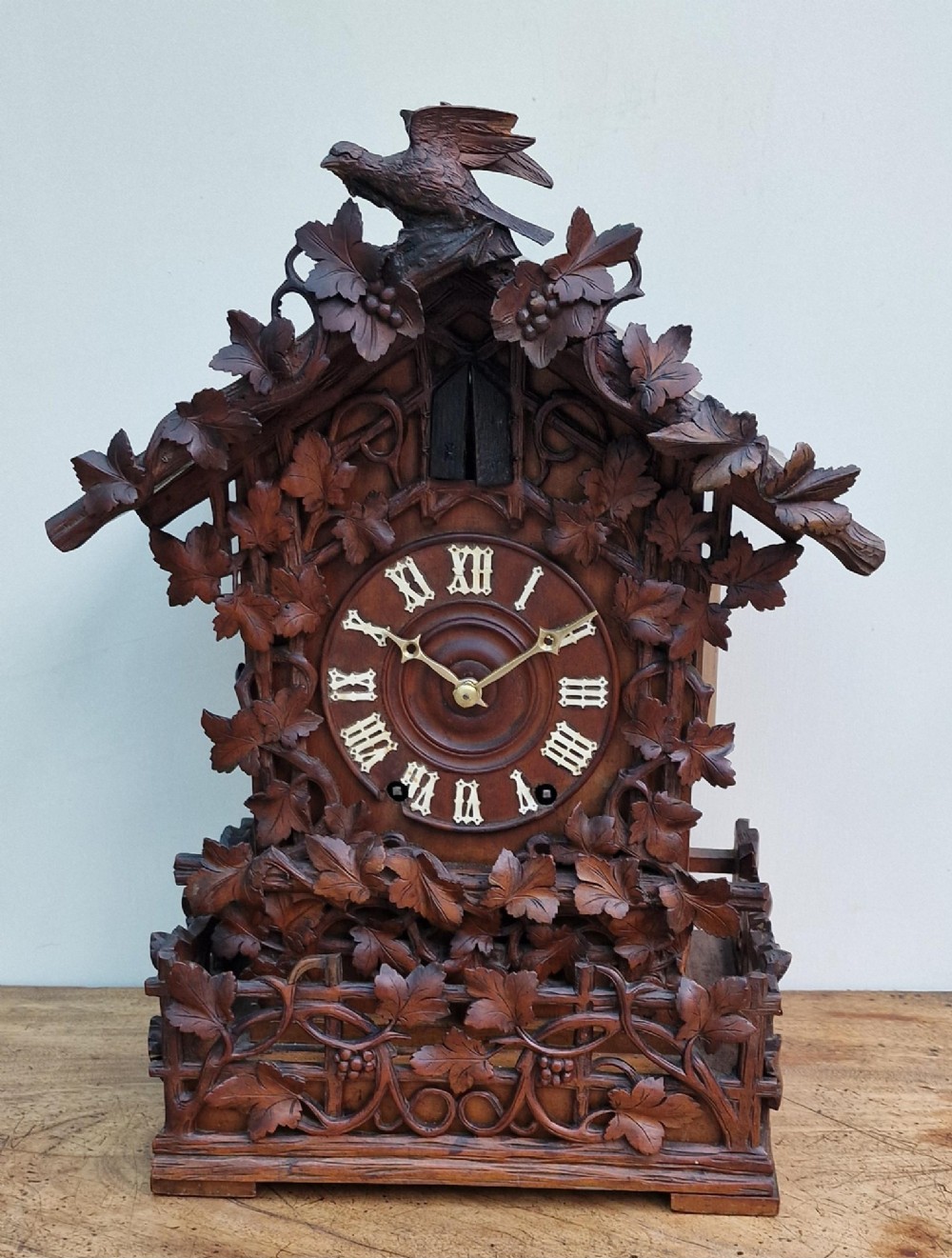 A dark brown carved wood cuckoo clock featuring intricate vine leaf motifs, a carved bird crest on the pitched roof, and Roman numerals on the circular dial face.