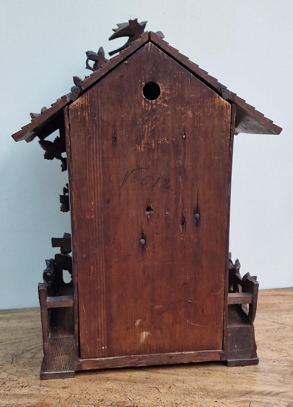A dark brown carved wood cuckoo clock featuring intricate vine leaf motifs, a carved bird crest on the pitched roof, and Roman numerals on the circular dial face. View II