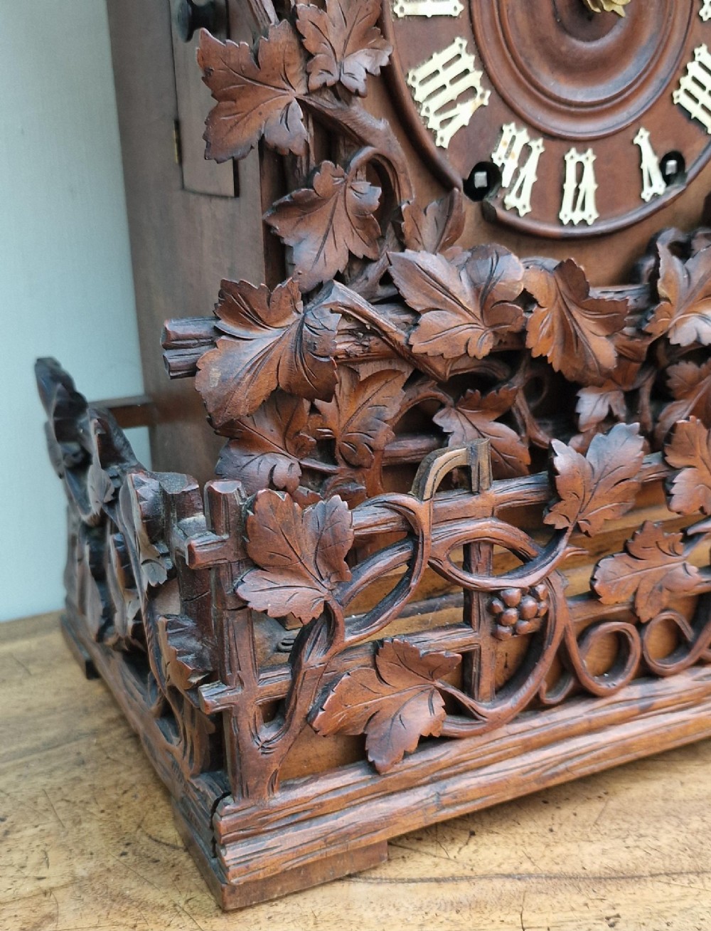 A dark brown carved wood cuckoo clock featuring intricate vine leaf motifs, a carved bird crest on the pitched roof, and Roman numerals on the circular dial face. View III