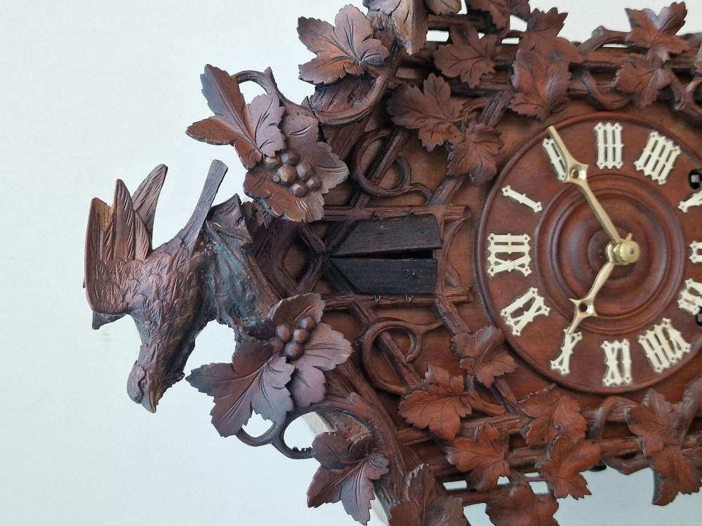 A dark brown carved wood cuckoo clock featuring intricate vine leaf motifs, a carved bird crest on the pitched roof, and Roman numerals on the circular dial face. View IV