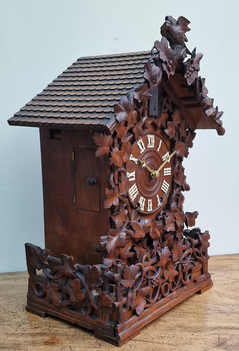 A dark brown carved wood cuckoo clock featuring intricate vine leaf motifs, a carved bird crest on the pitched roof, and Roman numerals on the circular dial face. View VII