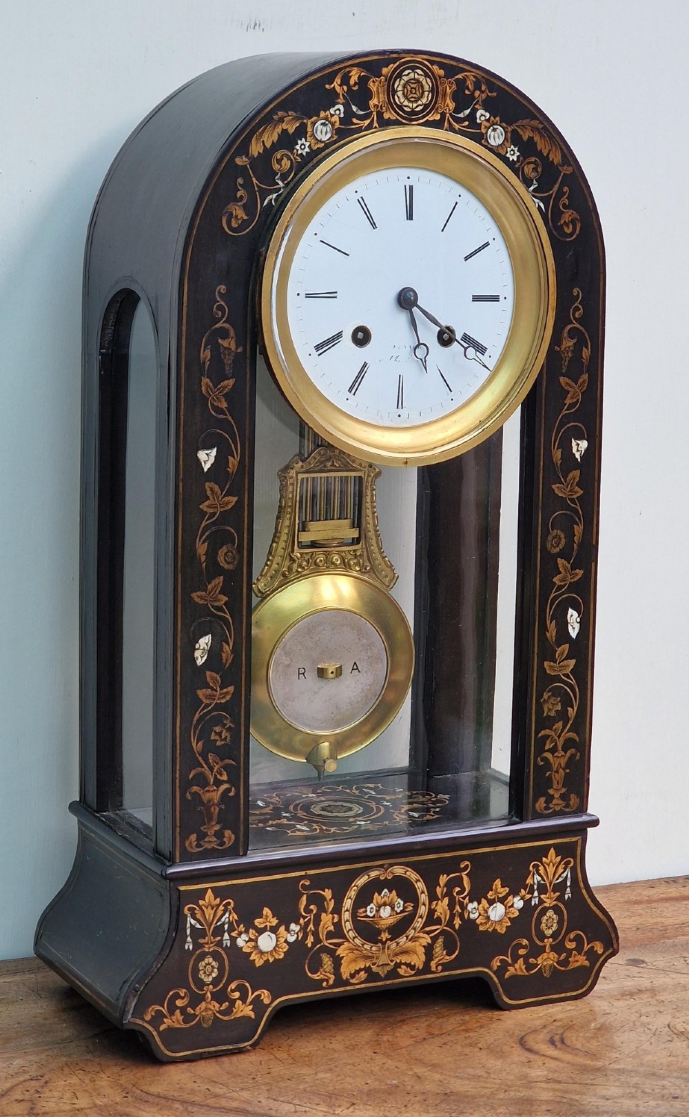An ebonised wooden mantel clock featuring floral brass and mother of pearl marquetry inlay, an arched glass front displaying a gridiron pendulum, and a white enamel dial with Roman numerals.