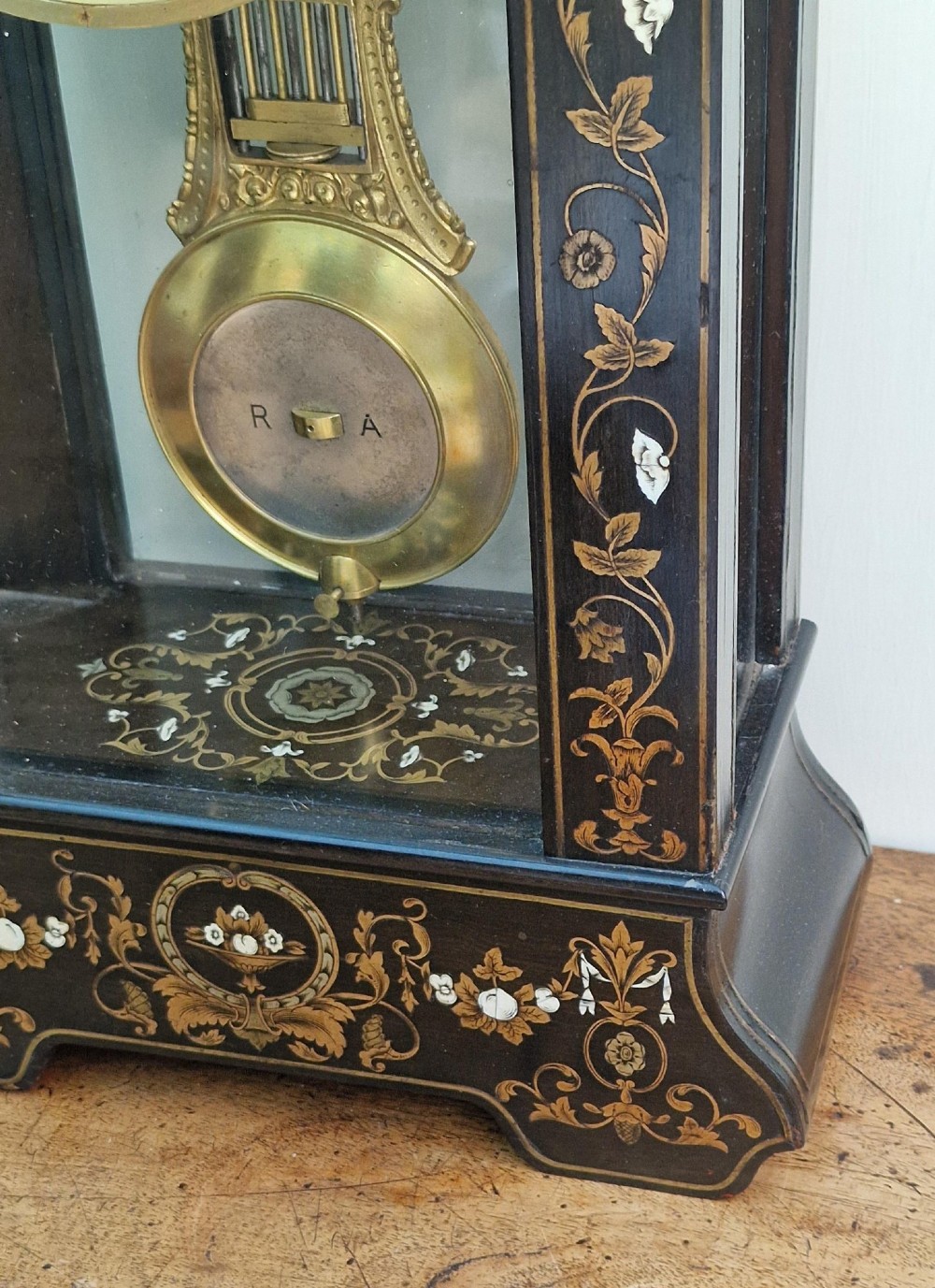 An ebonised wooden mantel clock featuring floral brass and mother of pearl marquetry inlay, an arched glass front displaying a gridiron pendulum, and a white enamel dial with Roman numerals. View II