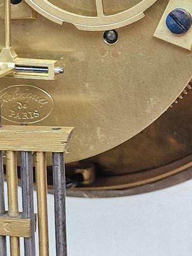 An ebonised wooden mantel clock featuring floral brass and mother of pearl marquetry inlay, an arched glass front displaying a gridiron pendulum, and a white enamel dial with Roman numerals. View V
