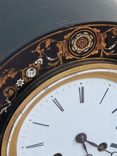 An ebonised wooden mantel clock featuring floral brass and mother of pearl marquetry inlay, an arched glass front displaying a gridiron pendulum, and a white enamel dial with Roman numerals. View VI