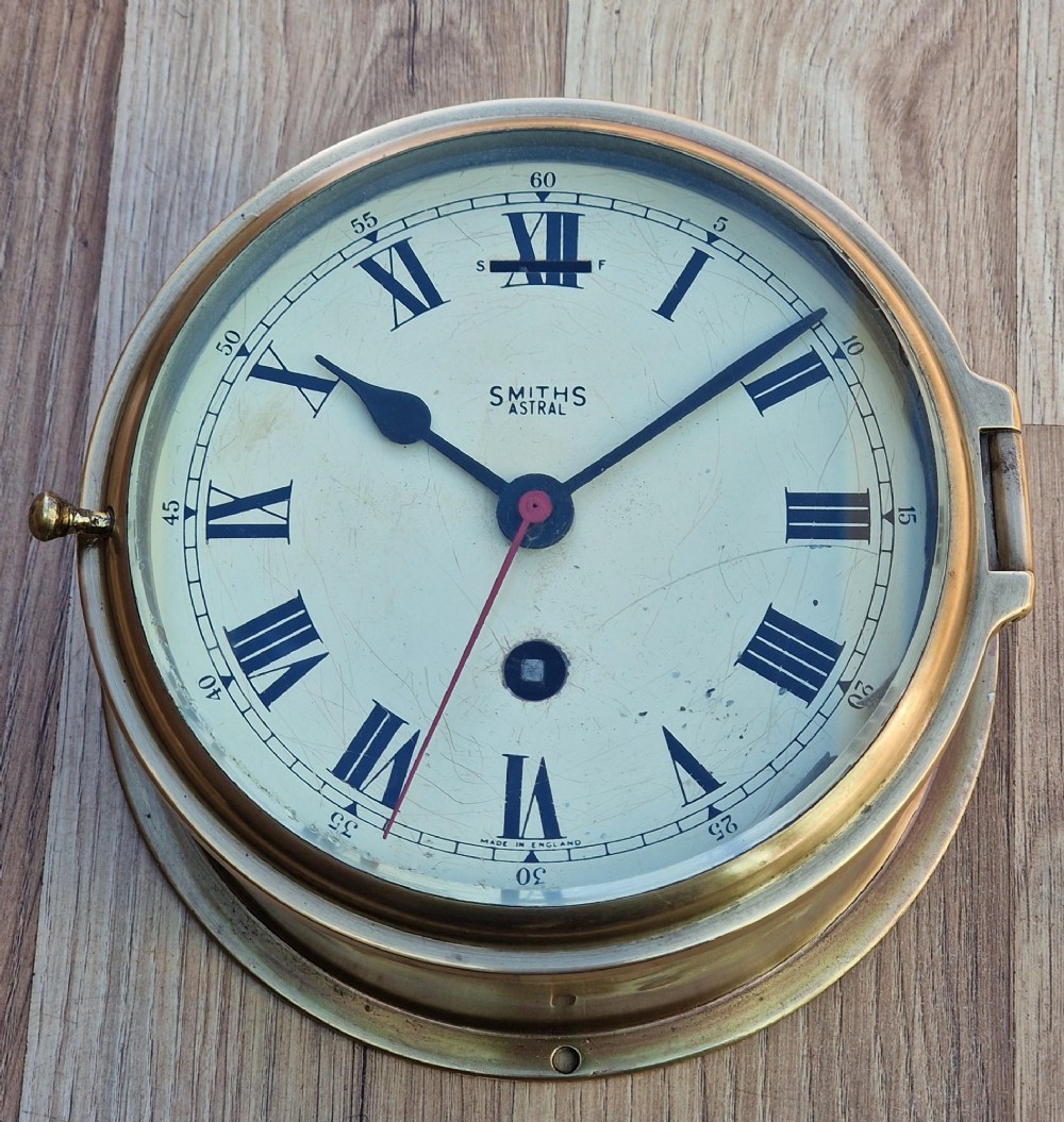A circular brass Smiths Astral ships bulkhead clock with a white face, black Roman numerals, and a red second hand.