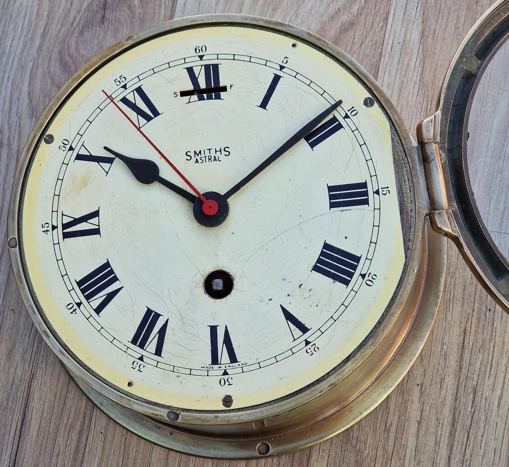 A circular brass Smiths Astral ships bulkhead clock with a white face, black Roman numerals, and a red second hand. View III