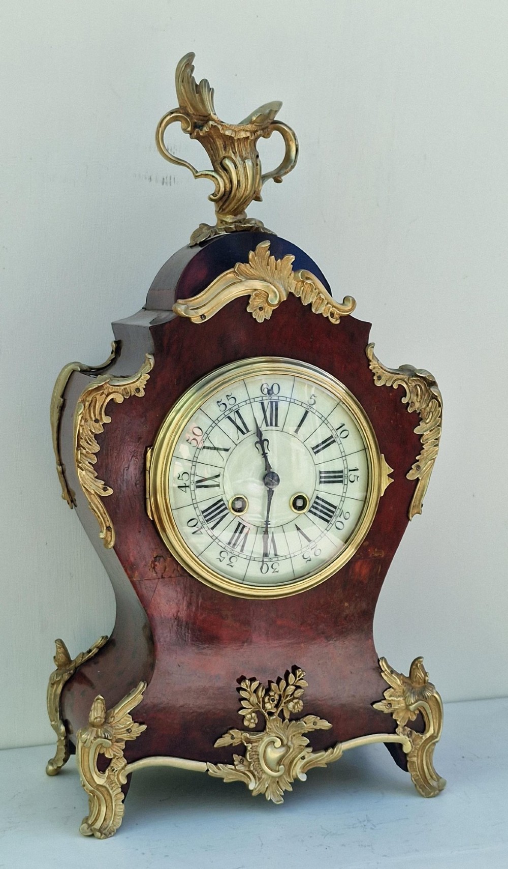 A French Rococo style mantel clock with a dark veneered balloon-shaped case, ornamental gilt-bronze foliage mounts, and a brass-rimmed dial showing Roman numerals.