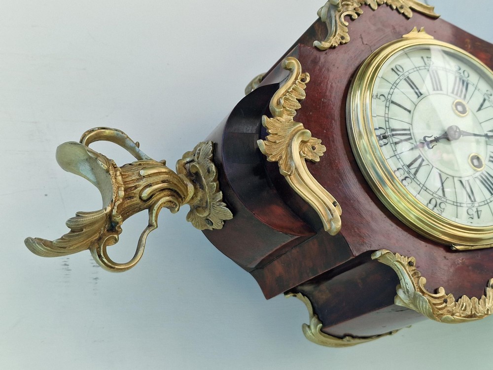A French Rococo style mantel clock with a dark veneered balloon-shaped case, ornamental gilt-bronze foliage mounts, and a brass-rimmed dial showing Roman numerals. View VI