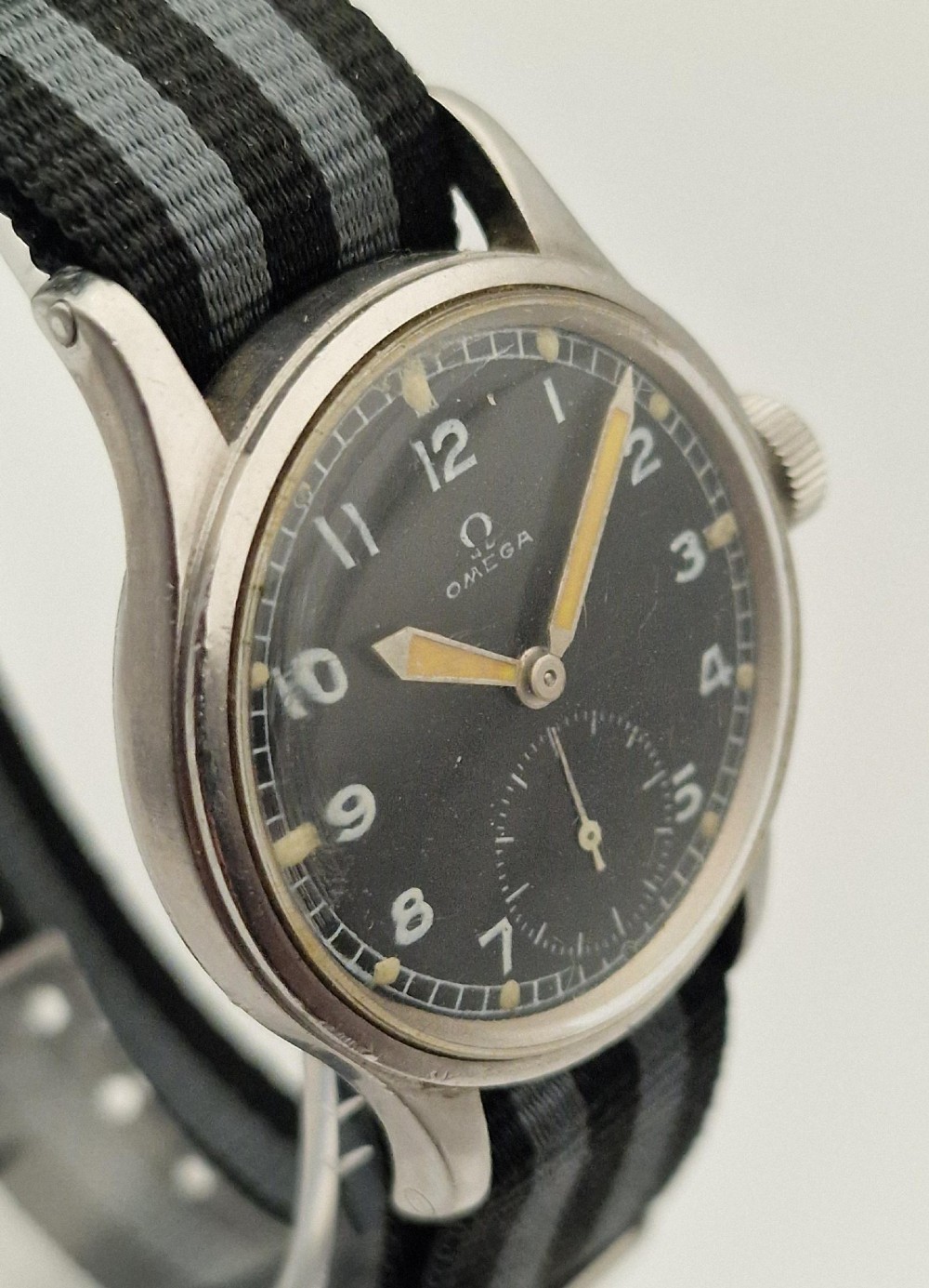A vintage 1945 Omega military wristwatch featuring a stainless steel case, black dial with luminous Arabic numerals, sub-seconds dial, and a black and grey striped fabric strap. View III