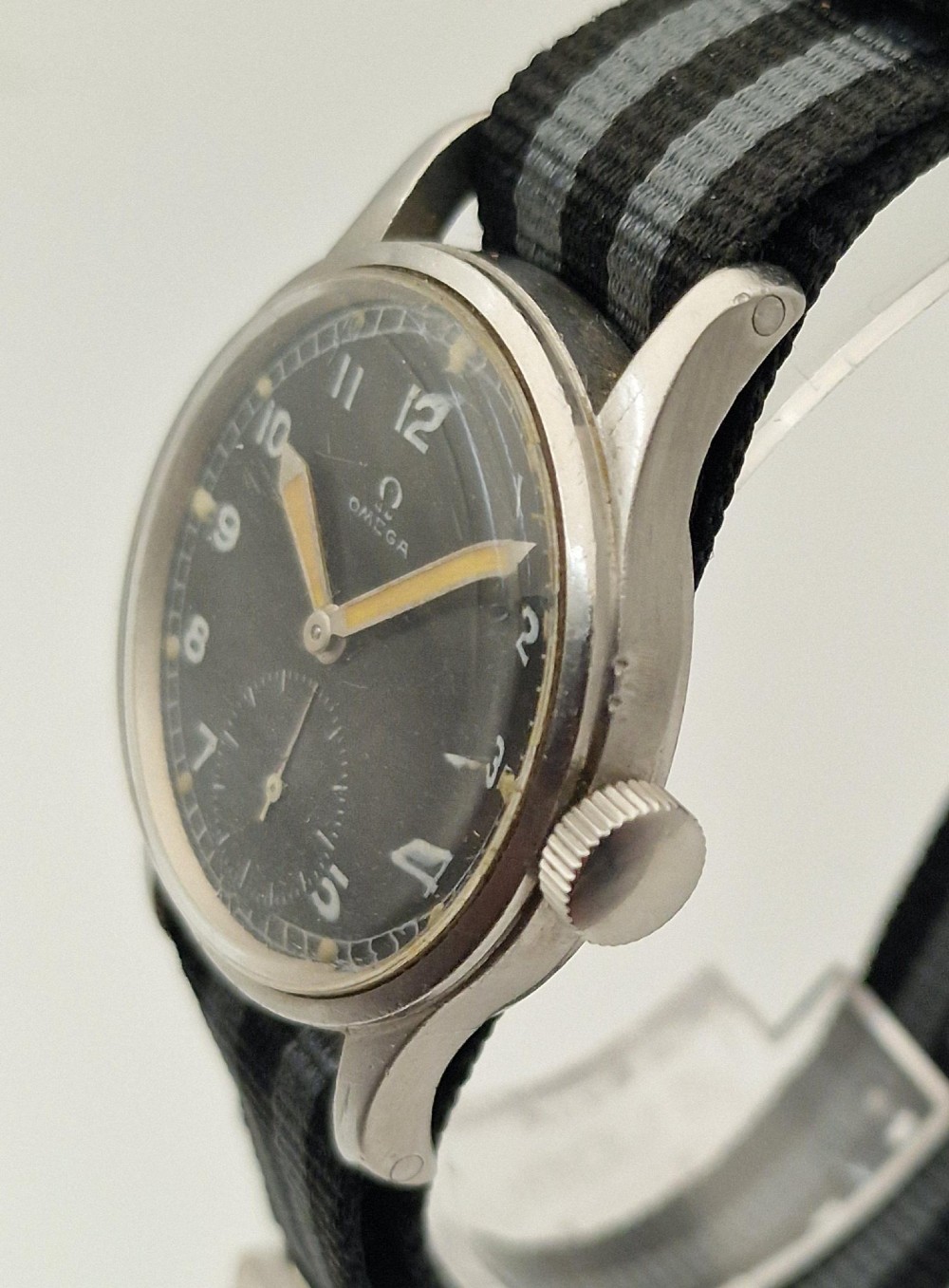 A vintage 1945 Omega military wristwatch featuring a stainless steel case, black dial with luminous Arabic numerals, sub-seconds dial, and a black and grey striped fabric strap. View VII