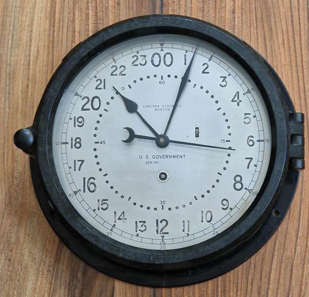 A circular black maritime wall clock featuring a twenty-four-hour dial face marked U.S. Government and Chelsea Clock Co. Boston.