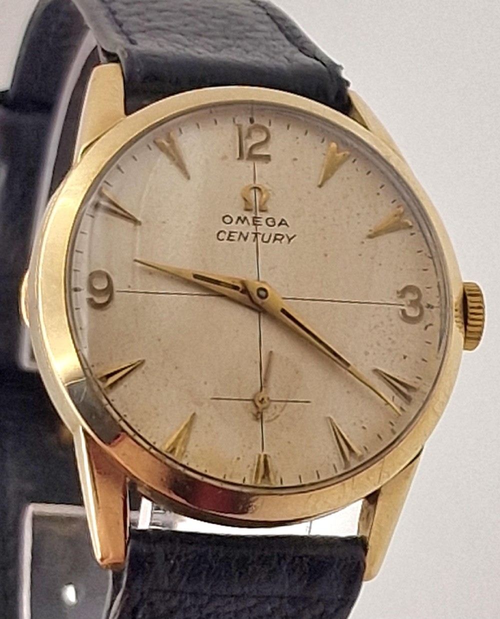 A 1954 Omega Century wristwatch with a gold-filled circular case, patinated dial with arrow markers, sub-seconds display, and a black leather strap.
