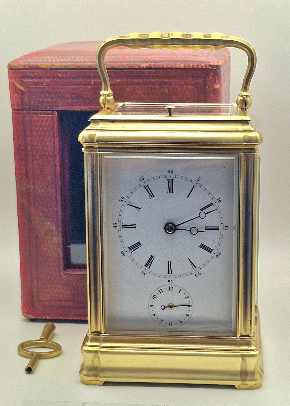 A brass carriage clock featuring a gorge style case, rectangular glass panels, a top carrying handle, a white enamel dial with Roman numerals, an accompanying key, and a red fitted travel case.