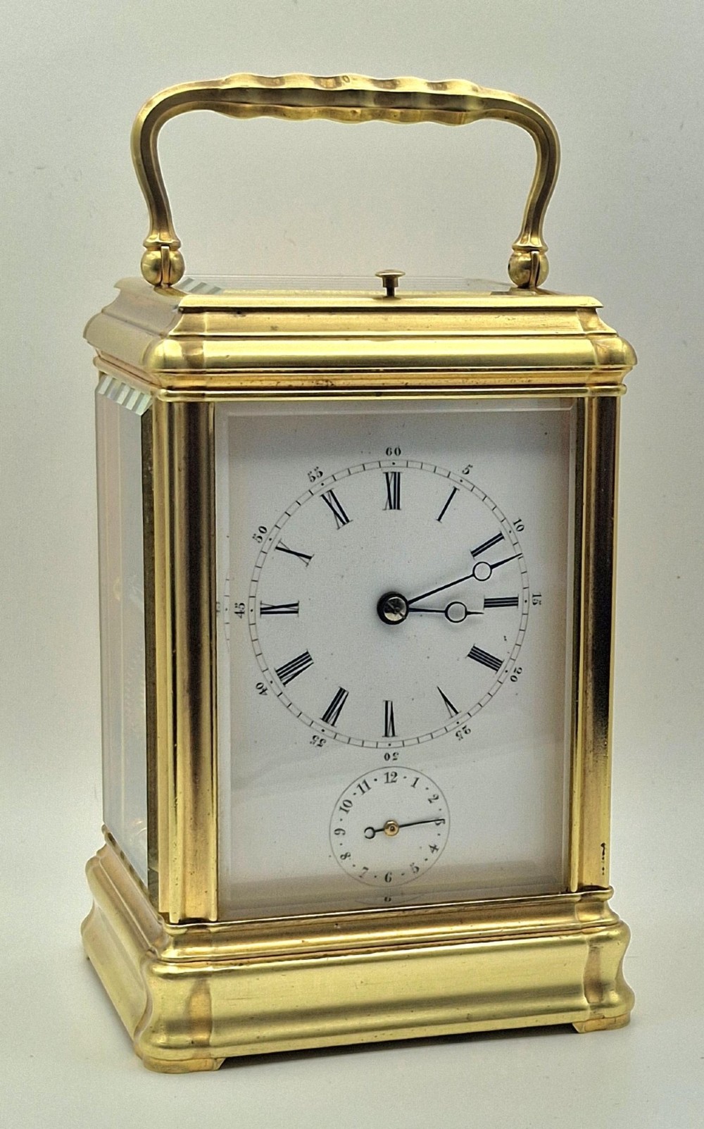 A brass carriage clock featuring a gorge style case, rectangular glass panels, a top carrying handle, a white enamel dial with Roman numerals, an accompanying key, and a red fitted travel case. View II