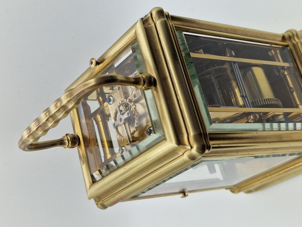 A brass carriage clock featuring a gorge style case, rectangular glass panels, a top carrying handle, a white enamel dial with Roman numerals, an accompanying key, and a red fitted travel case. View III