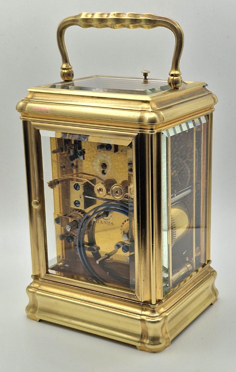 A brass carriage clock featuring a gorge style case, rectangular glass panels, a top carrying handle, a white enamel dial with Roman numerals, an accompanying key, and a red fitted travel case. View IV