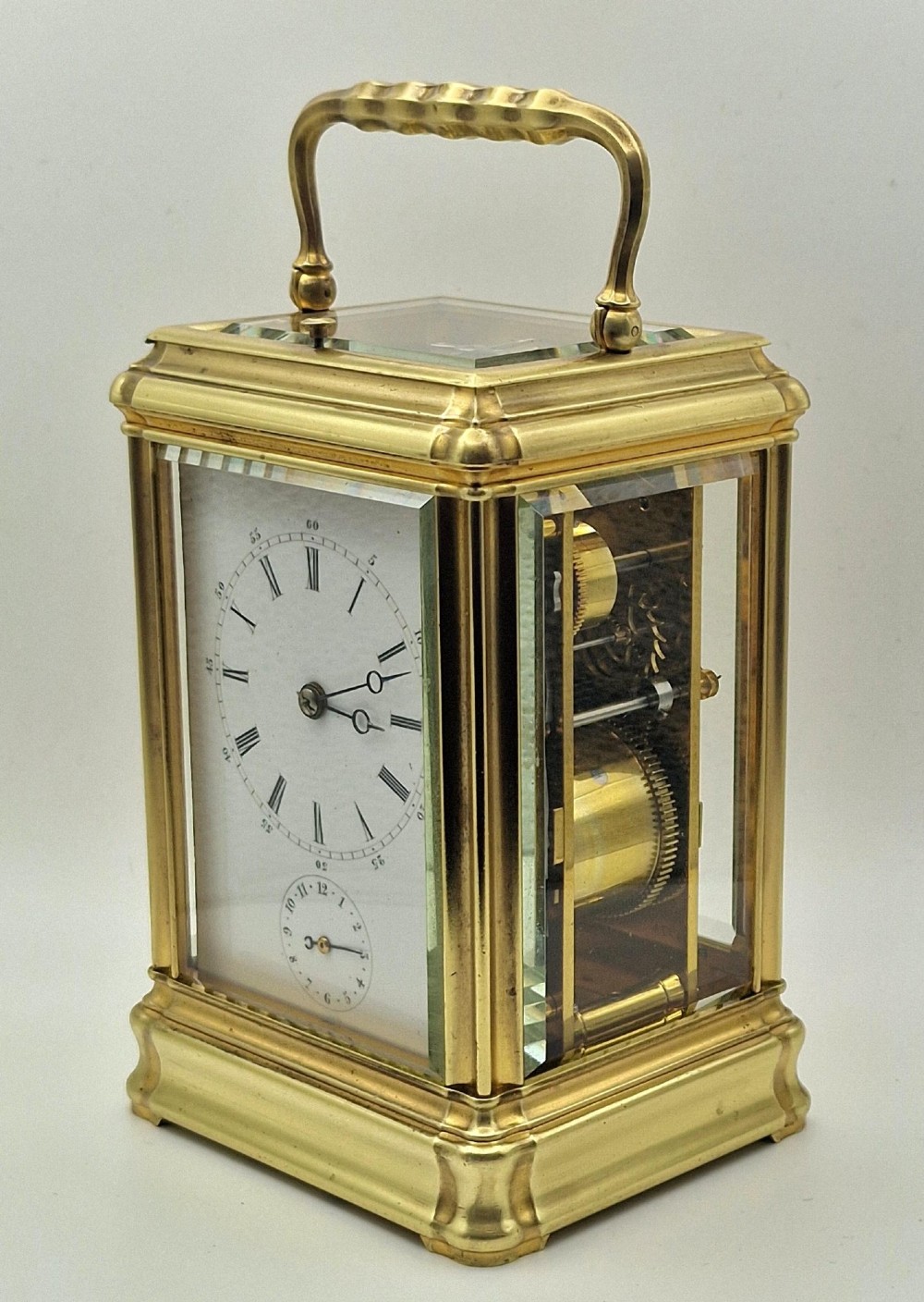 A brass carriage clock featuring a gorge style case, rectangular glass panels, a top carrying handle, a white enamel dial with Roman numerals, an accompanying key, and a red fitted travel case. View V
