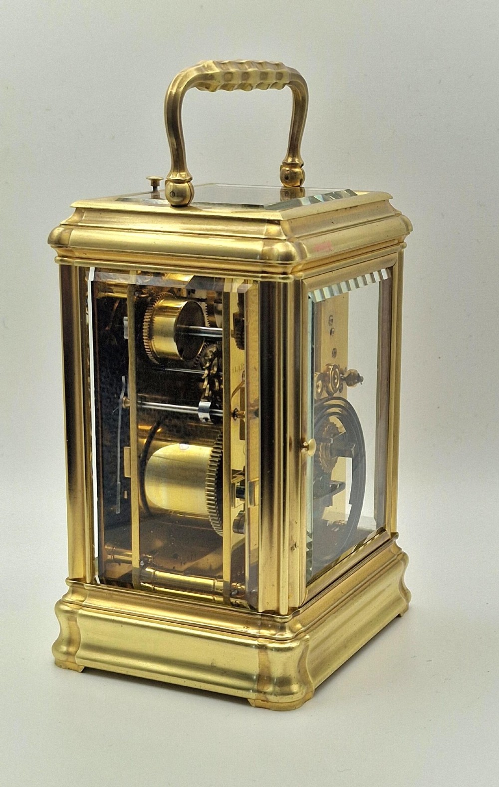 A brass carriage clock featuring a gorge style case, rectangular glass panels, a top carrying handle, a white enamel dial with Roman numerals, an accompanying key, and a red fitted travel case. View VI