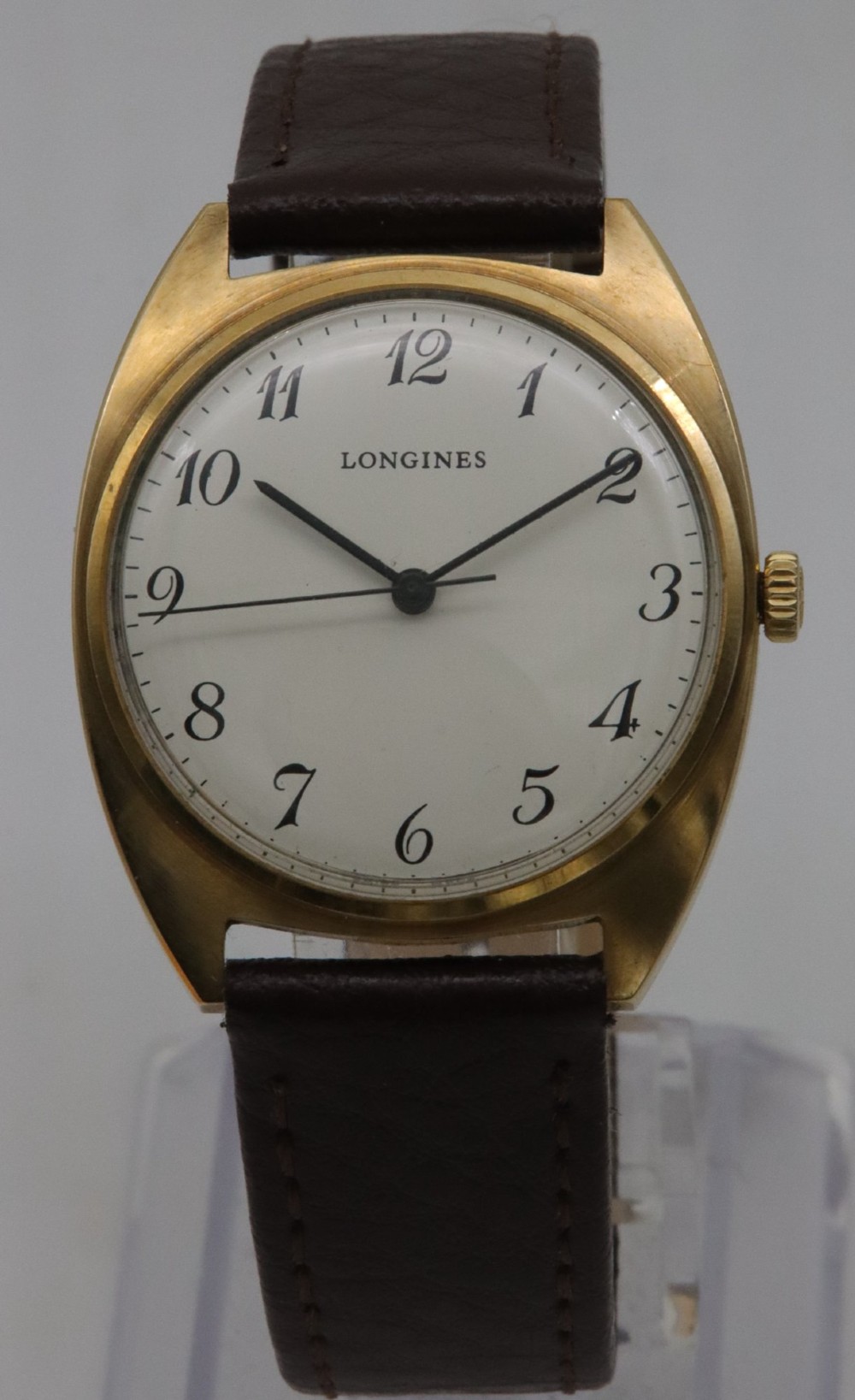 A 1974 Longines wristwatch with a cushion-shaped 9k yellow gold case, a white dial with black Arabic numerals, and a brown leather strap.