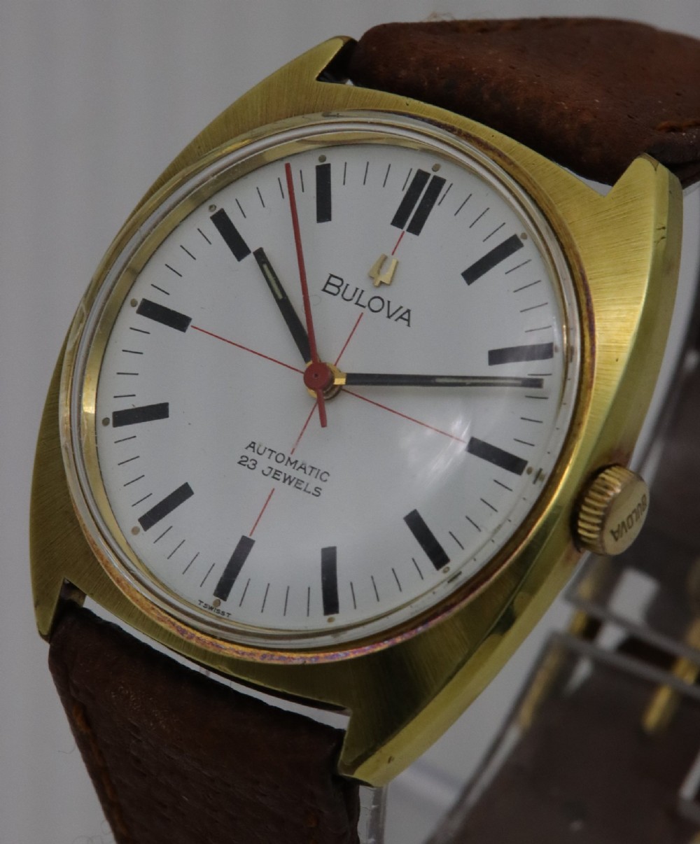 A 1970s Bulova automatic wristwatch featuring a gold-toned cushion case, a white crosshair dial with black markers, and a brown leather strap. View II