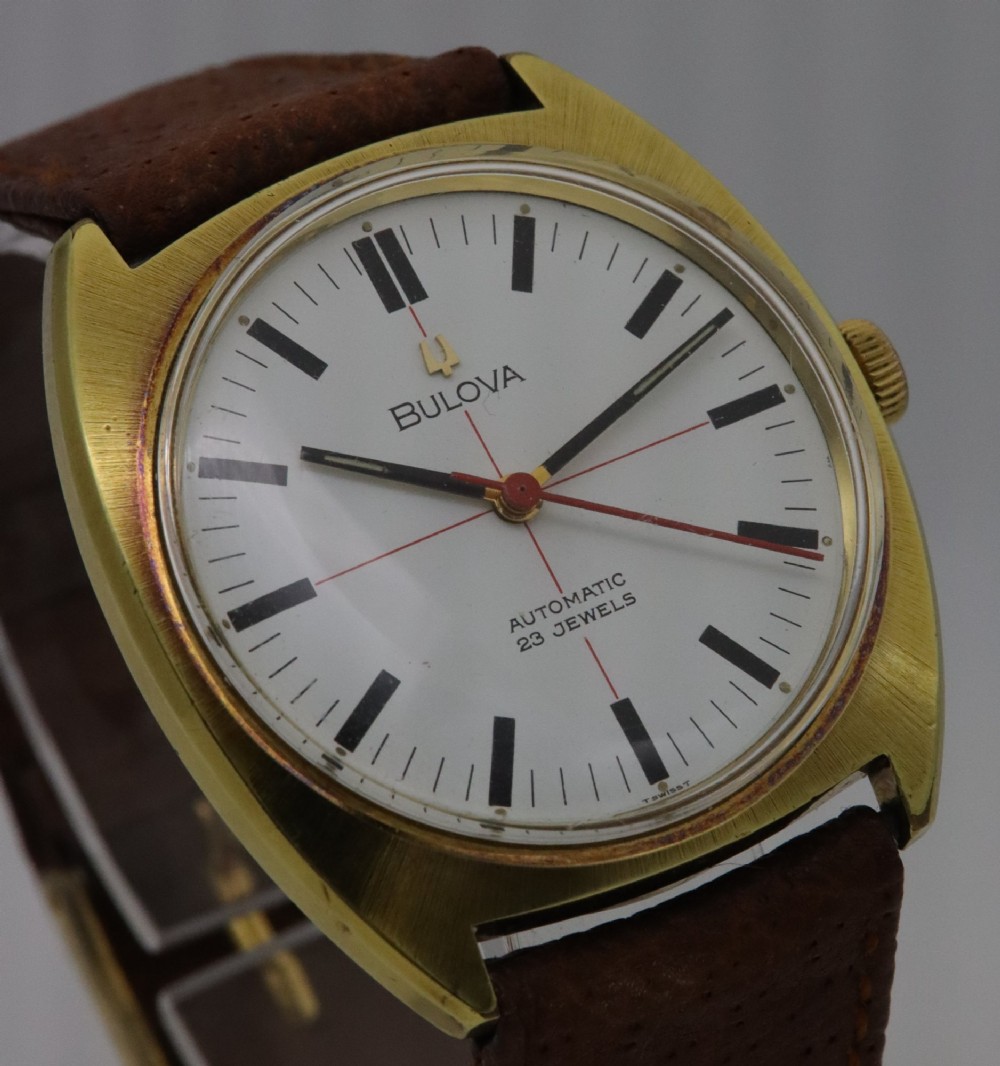 A 1970s Bulova automatic wristwatch featuring a gold-toned cushion case, a white crosshair dial with black markers, and a brown leather strap. View IV