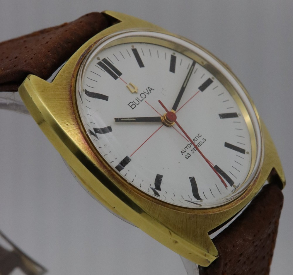 A 1970s Bulova automatic wristwatch featuring a gold-toned cushion case, a white crosshair dial with black markers, and a brown leather strap. View V