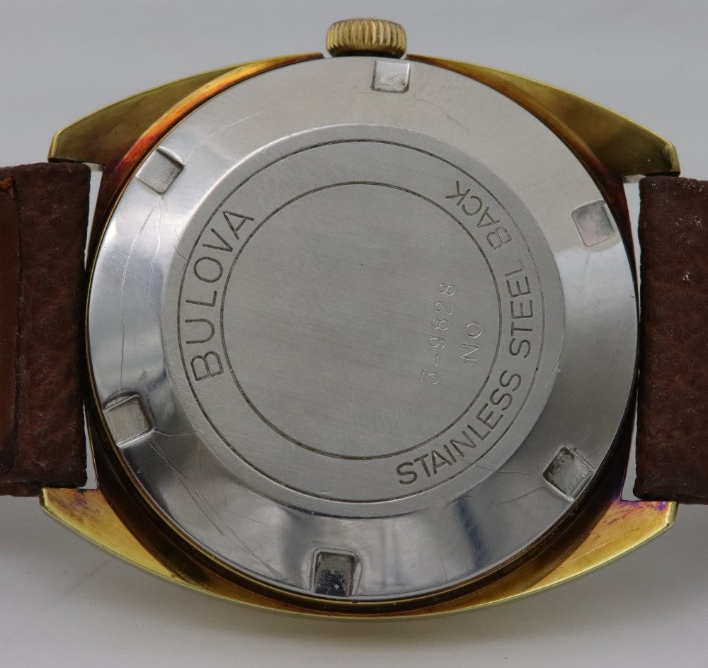A 1970s Bulova automatic wristwatch featuring a gold-toned cushion case, a white crosshair dial with black markers, and a brown leather strap. View VI