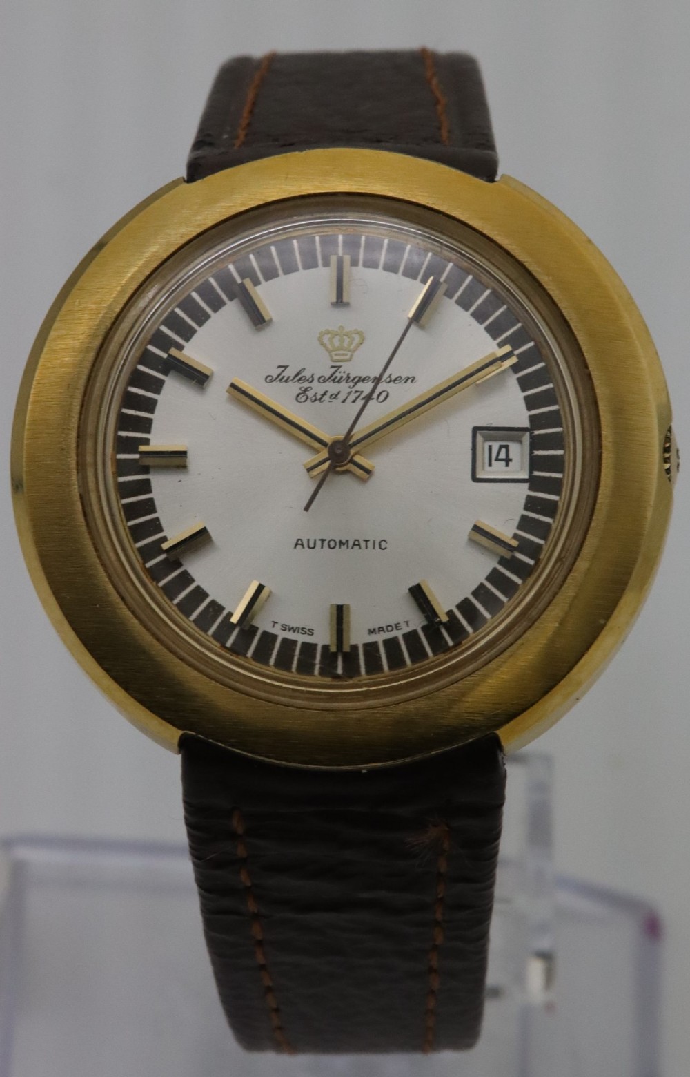 A 1970s Jules Jurgensen automatic wristwatch with a gold-toned circular cushion case, white dial, date aperture, and brown textured leather strap.
