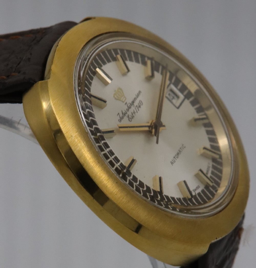 A 1970s Jules Jurgensen automatic wristwatch with a gold-toned circular cushion case, white dial, date aperture, and brown textured leather strap. View V