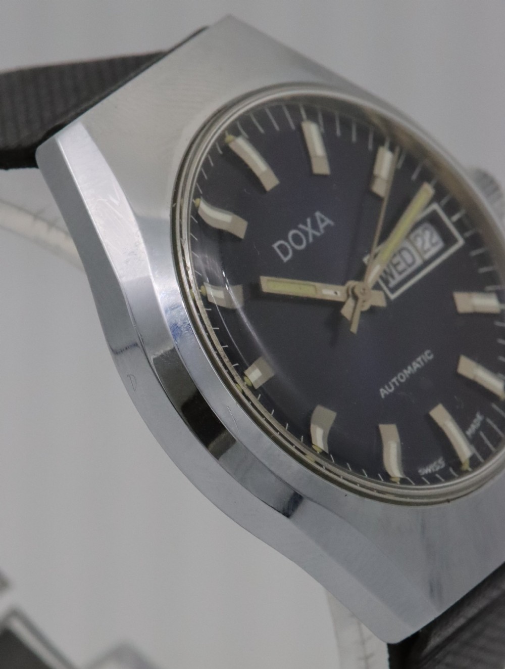 A 1970s Doxa automatic wristwatch featuring a cushion-shaped stainless steel case, blue dial with day-date aperture, and black strap. View V
