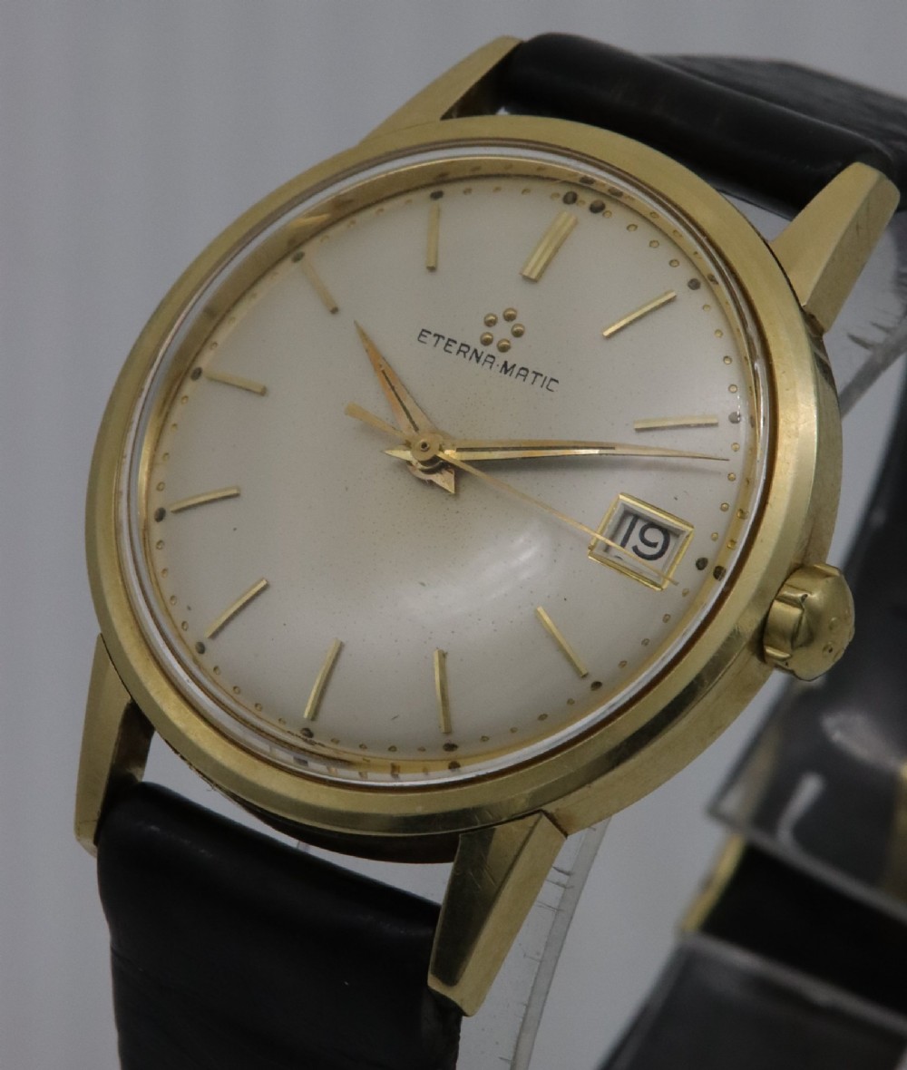 A 1957 Eterna-Matic 14K gold wristwatch with an off-white dial, date display window at three oclock, gold baton indices, and a black leather strap. View II