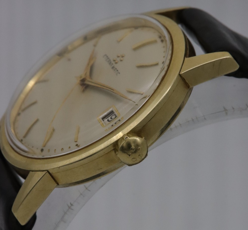 A 1957 Eterna-Matic 14K gold wristwatch with an off-white dial, date display window at three oclock, gold baton indices, and a black leather strap. View III