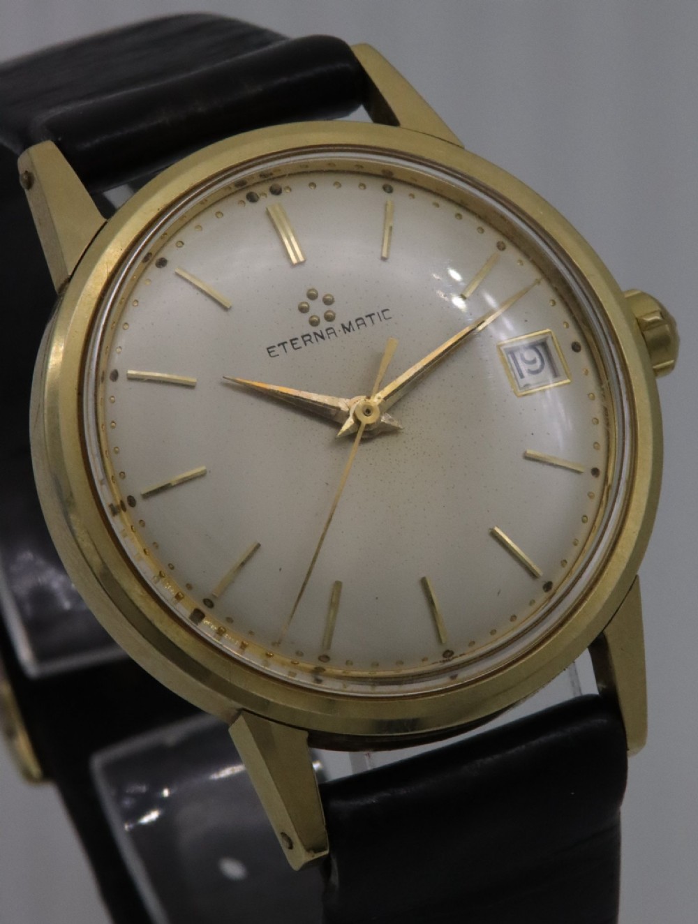 A 1957 Eterna-Matic 14K gold wristwatch with an off-white dial, date display window at three oclock, gold baton indices, and a black leather strap. View IV