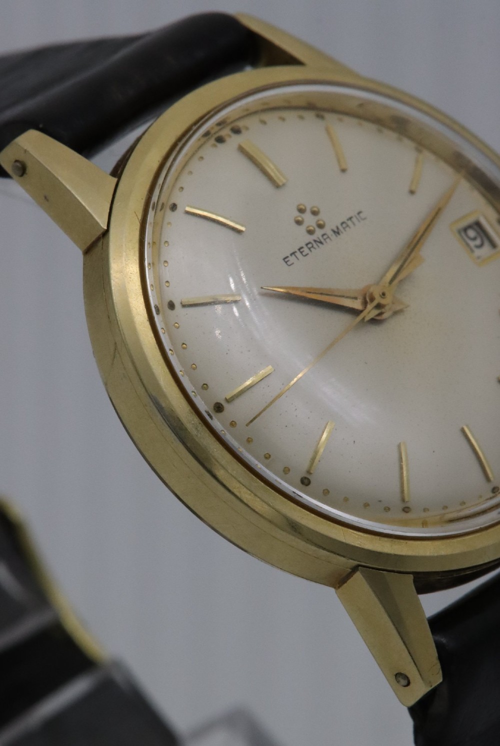 A 1957 Eterna-Matic 14K gold wristwatch with an off-white dial, date display window at three oclock, gold baton indices, and a black leather strap. View V