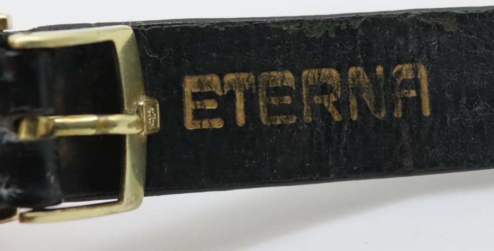 A 1957 Eterna-Matic 14K gold wristwatch with an off-white dial, date display window at three oclock, gold baton indices, and a black leather strap. View VI