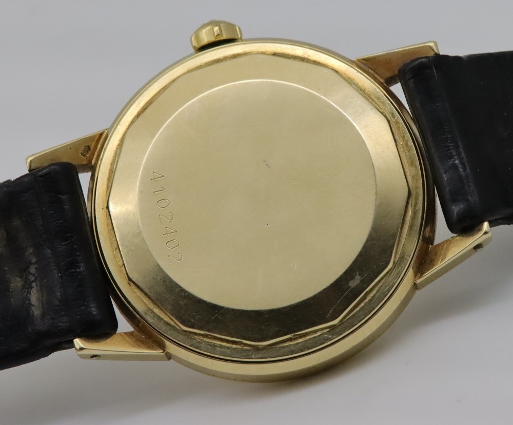 A 1957 Eterna-Matic 14K gold wristwatch with an off-white dial, date display window at three oclock, gold baton indices, and a black leather strap. View VII