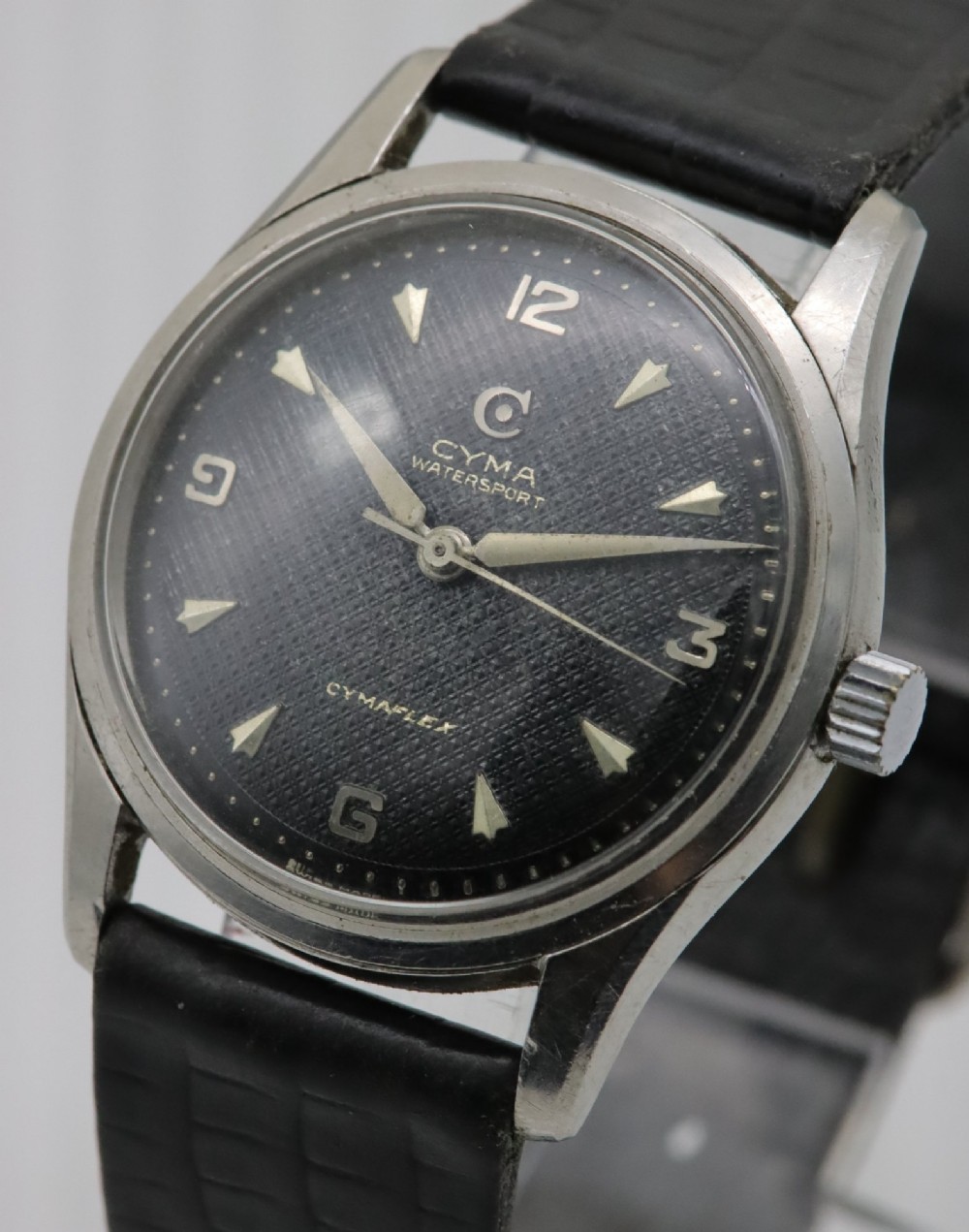 A 1950s Cyma Watersport Cymaflex wristwatch featuring a round stainless steel case, a black textured waffle dial with silver markers, and a black strap. View II