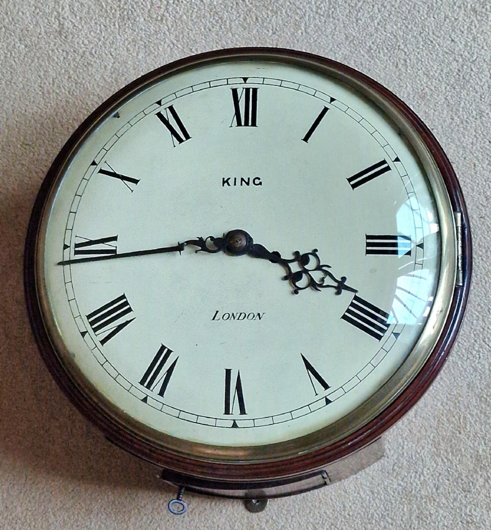 A circular Georgian fusee wall clock with a dark mahogany bezel surround, off-white painted dial featuring Roman numerals, ornate black hands, and signed King London.