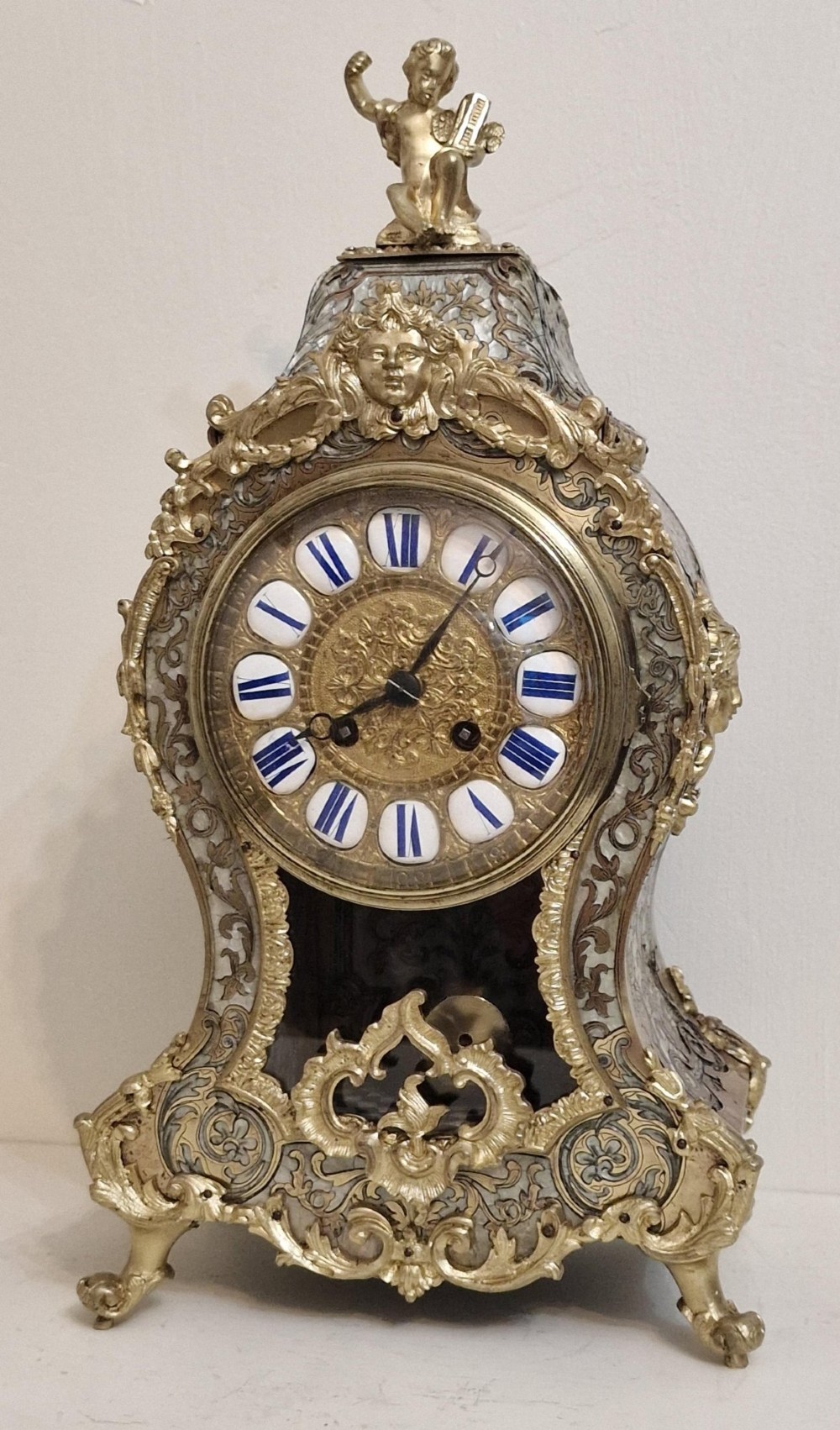 A French Rococo revival Boulle inlaid mantel clock featuring gilt brass mounts, blue Roman numerals on white enamel cartouches, and a cherub finial.
