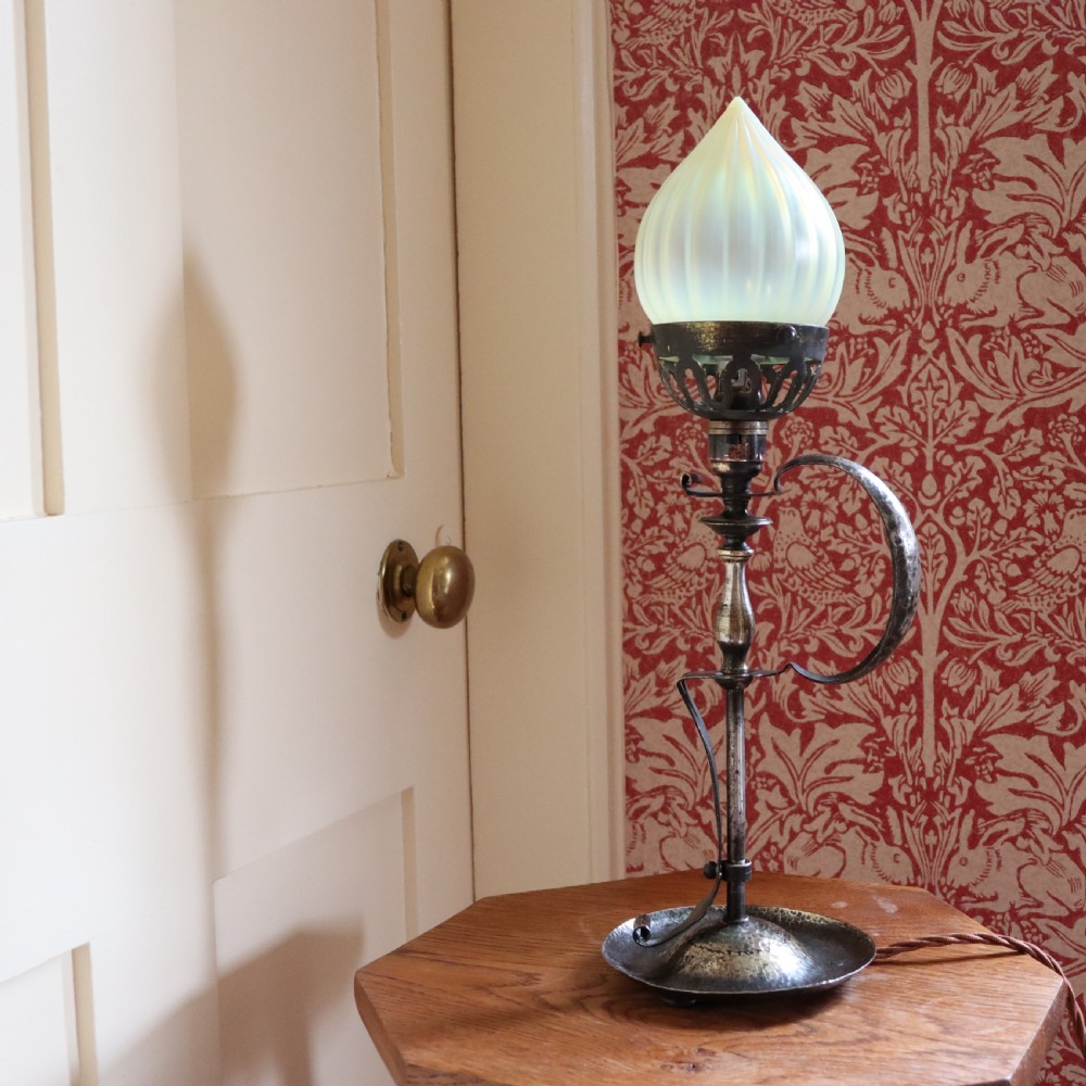 An Arts and Crafts brass table lamp featuring a hand-hammered circular base, a curved handle stem, and a flame-shaped ribbed vaseline glass shade.